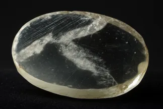 Lente de Nimrud realista sobre fondo negro de estudio; cristal de roca ovalado con rayas de pulido neoasirrias.