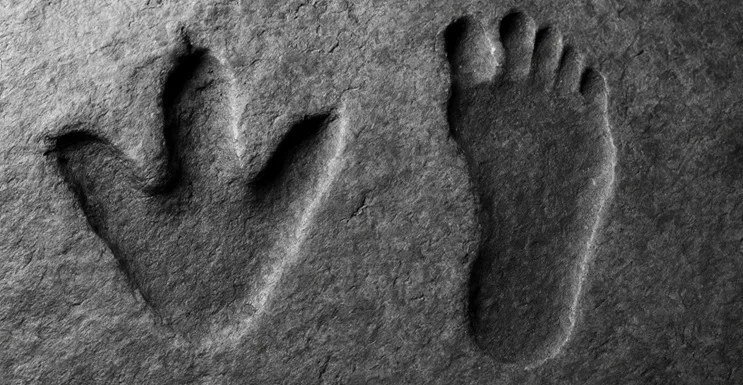 Huellas fósiles de dinosaurio y huella humana lado a lado en piedra arenisca, iluminadas lateralmente